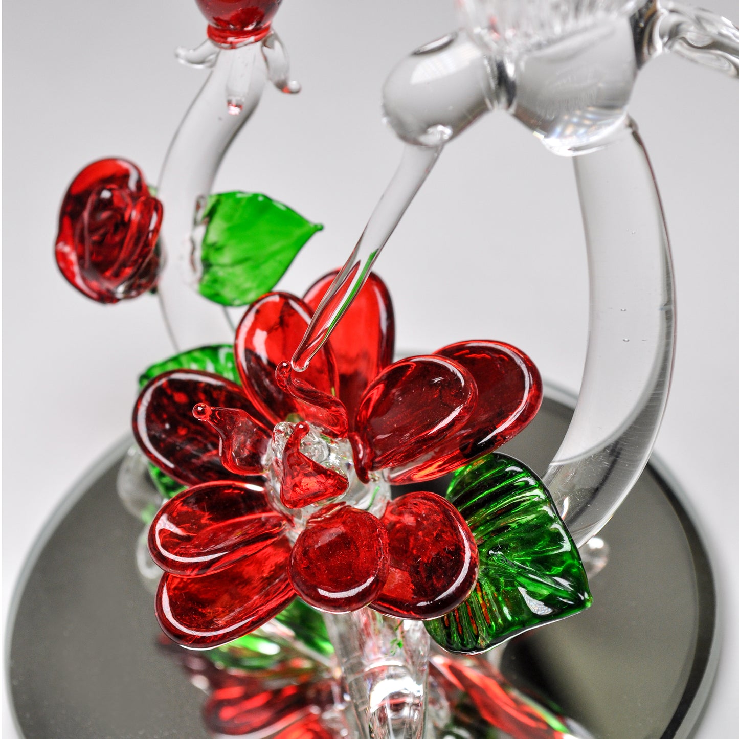 Crystal Castle Hummingbird flying over red and green glass flowers and flowers buds on a mirror base. Close up of biggest blooming glass blown rose.